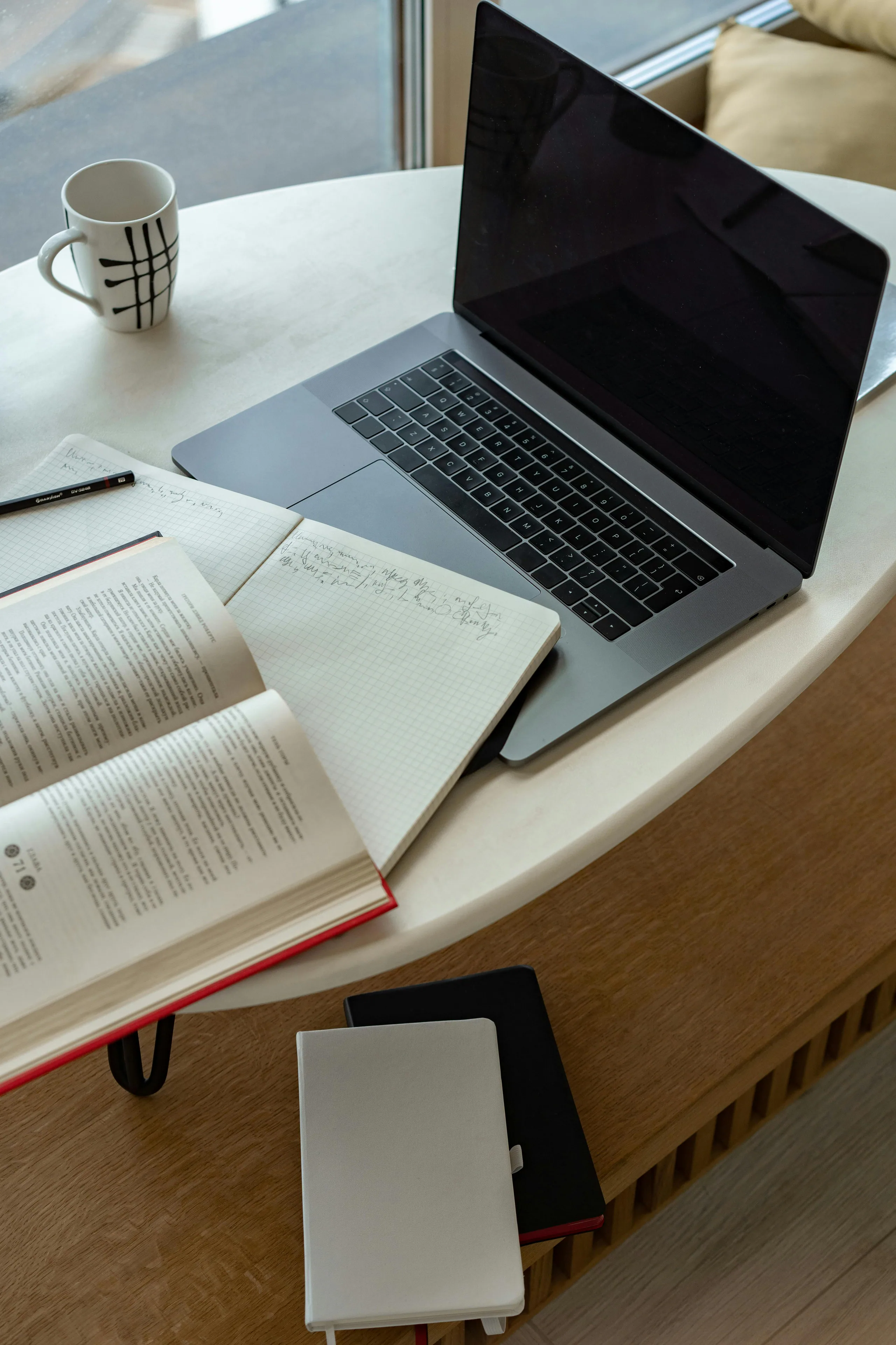 Open schriftje met notities, een boek, een laptop en een koffietas op een tafel